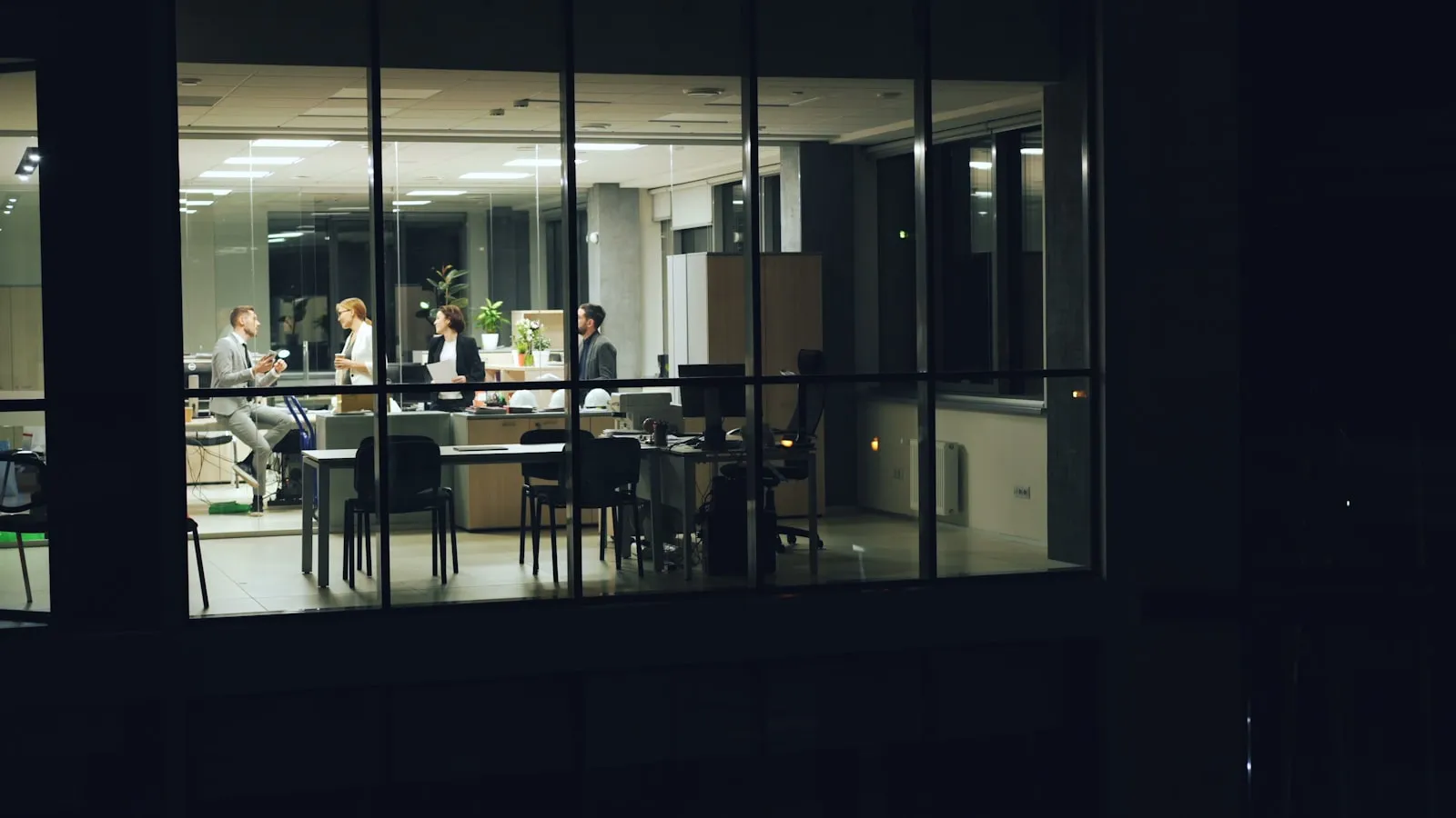 Vue nocturne à travers la façade vitrée d'un plateau de bureaux, où quelques professionnels échangent debout.